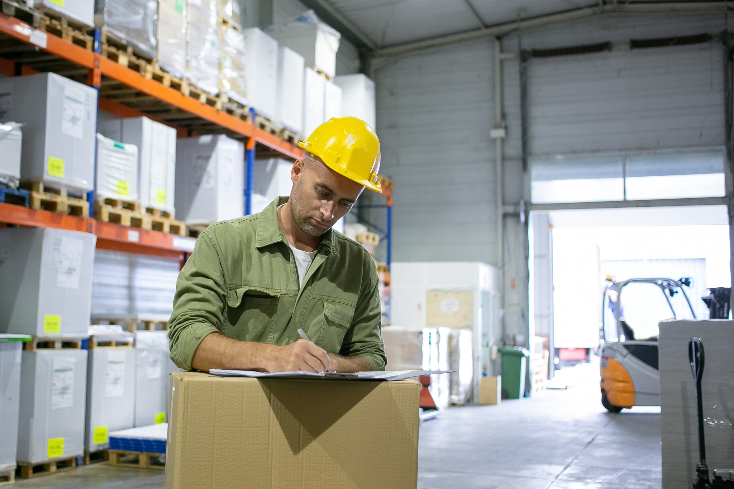 serious male logistic engineer in hardhat and overall standing at stack of boxes and completing documents copy space blue collar or labor concept 1 scaled