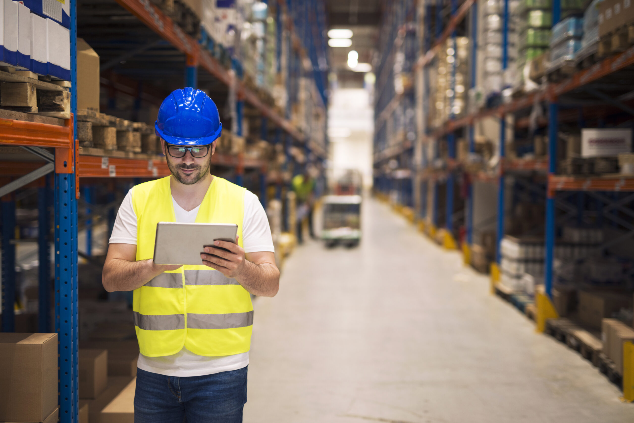 warehouse worker checking inventory on his tablet while walking in large storage department with shelves and packages in background scaled