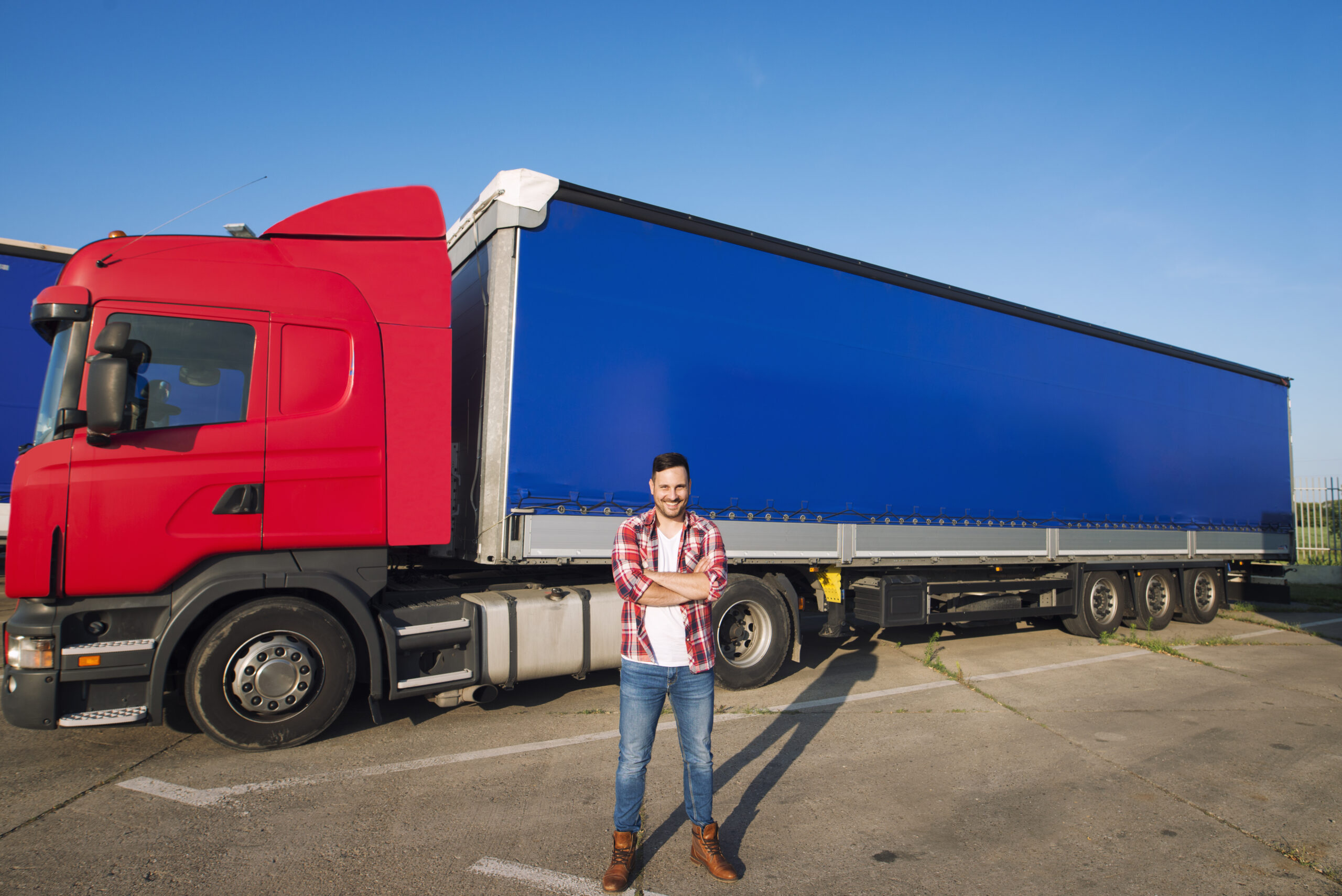 portrait of professional american truck driver in casual clothing and boots standing in front of truck vehicle with long trailer scaled